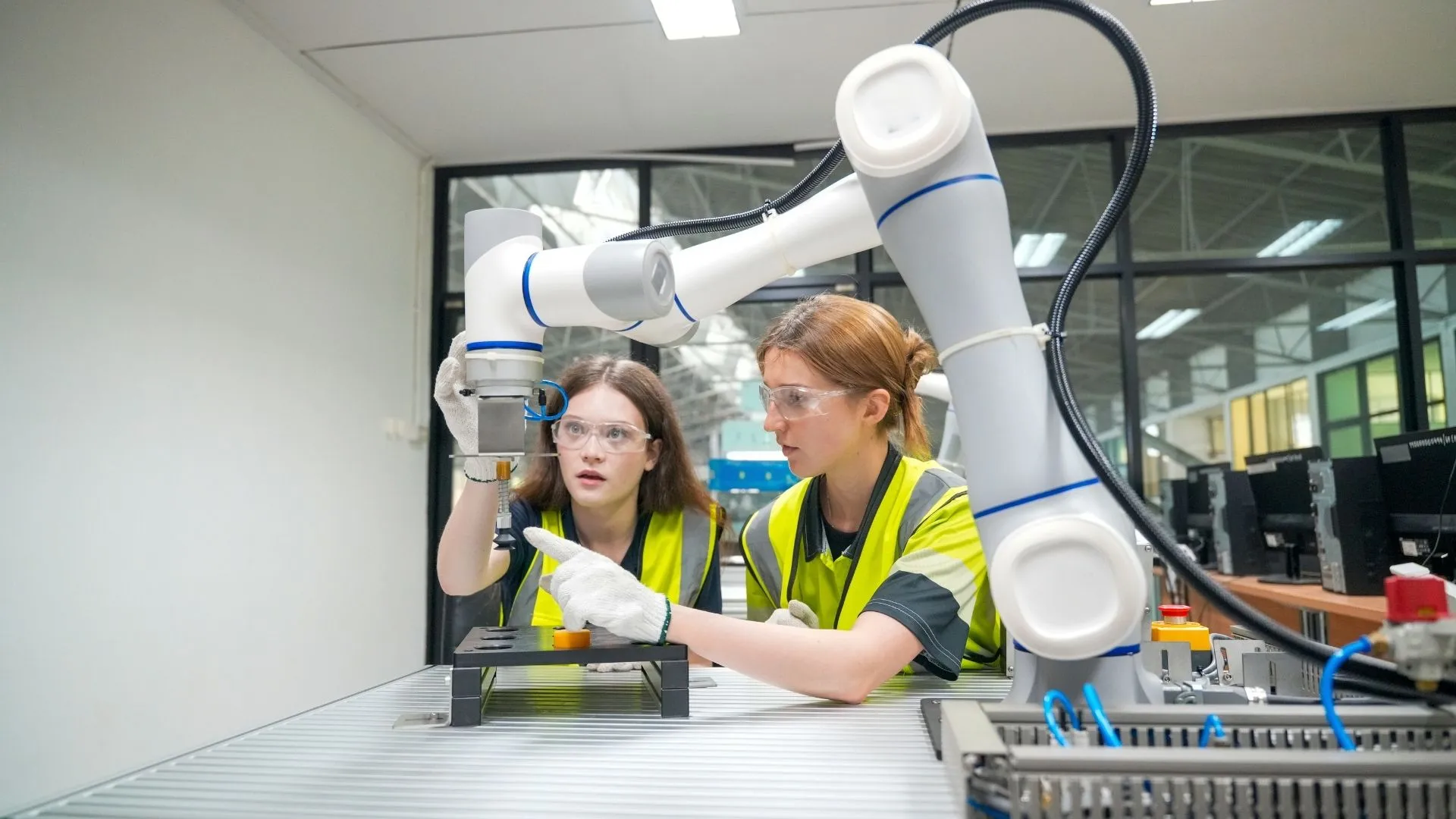 Deux apprenties en gilet jaune devant un bras robotisé dans un atelier