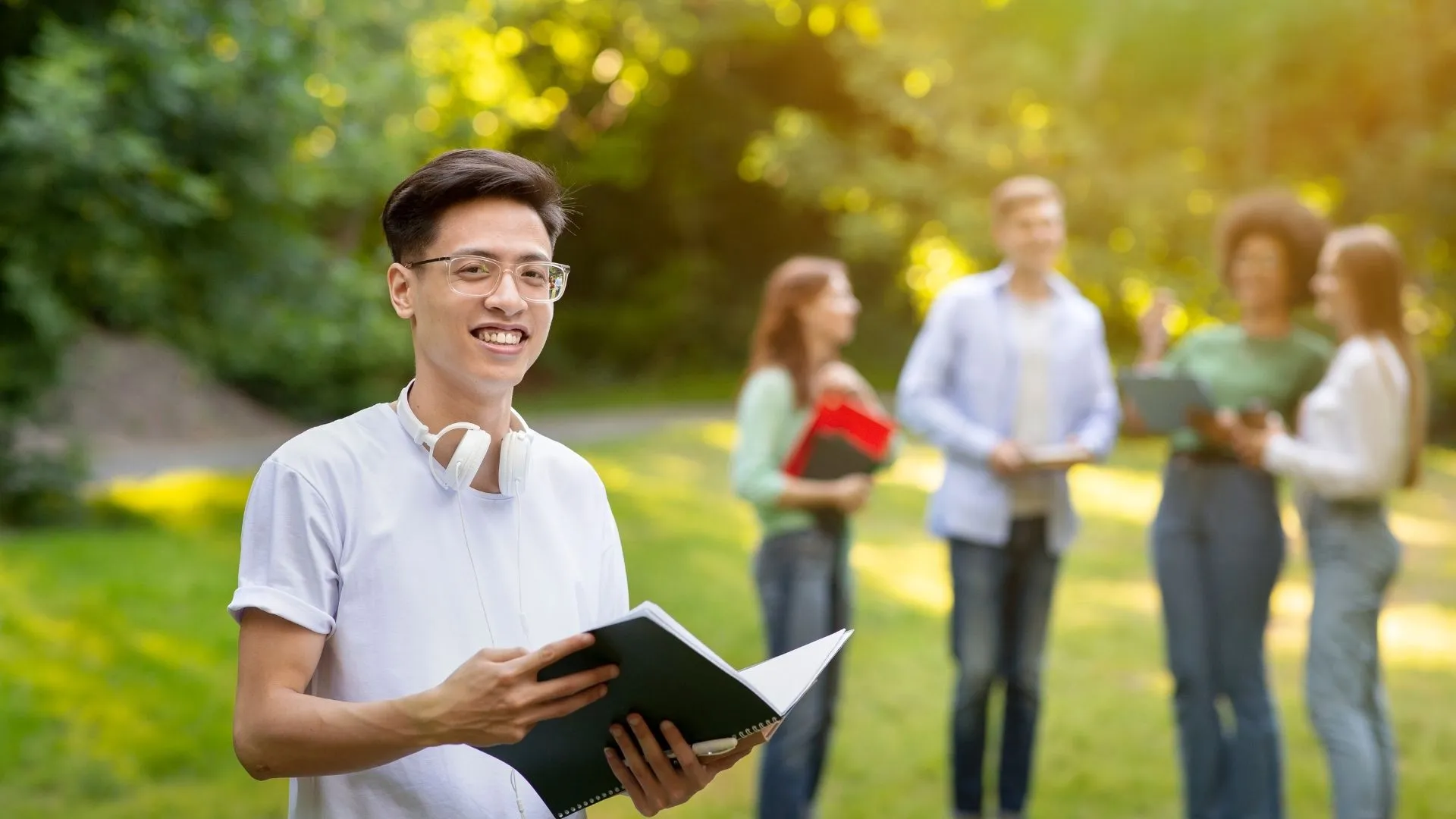 Un étudiant souriant tient un cahier sur un campus, des camarades discutent derrière lui